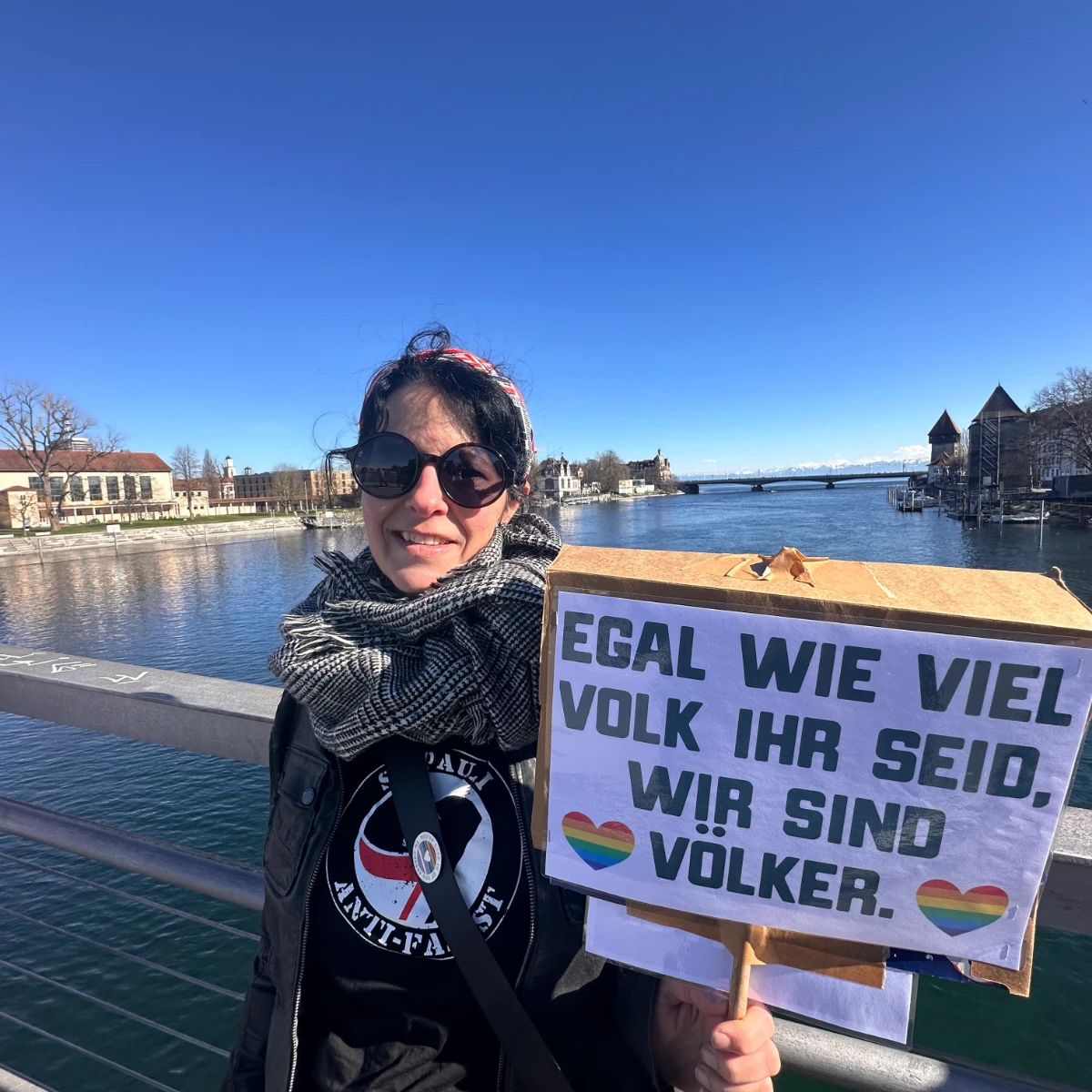 Eine Frau steht auf der Fahrradbrücke über dem Rhein in Konstanz und geht offensichtlich zu einer Demonstration. Sie trägt ein St. Pauli Antifa-Shirt, ein LGBTQ+ Button und hat ein Demoplakat in den Händen. Auf dem Plakat steht "Egal wie viel Volk ihr seid, wir sind Völker."